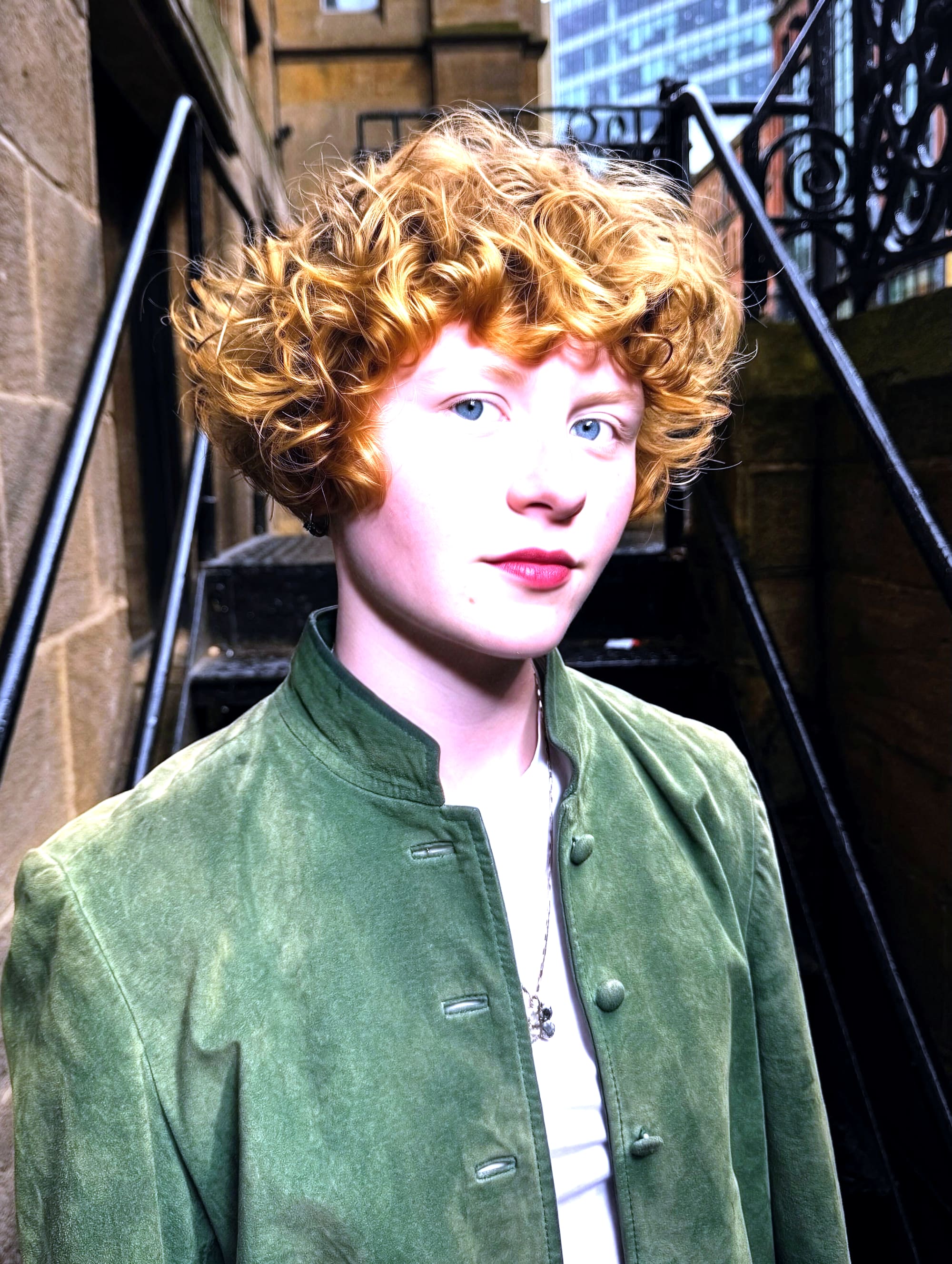 Copper curly crop and red lip on a wrought-iron staircase in Leeds — green suede jacket, direct gaze.